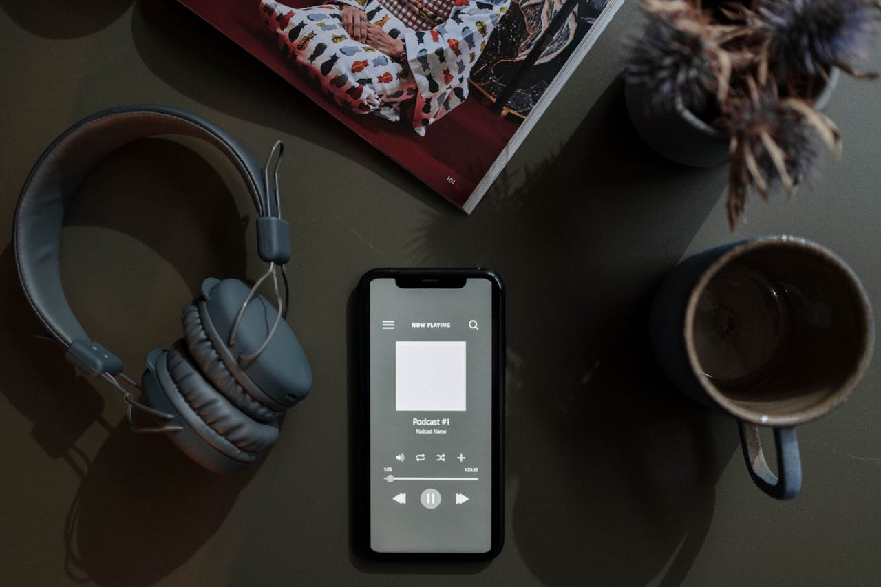 A flat lay image of a smartphone playing a podcast next to headphones and a mug.
